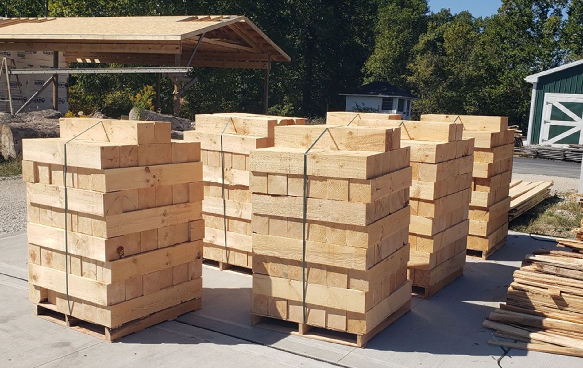 Bundles of stacked, light-colored wooden beams sit on pallets on a concrete outdoor lot near a construction site.