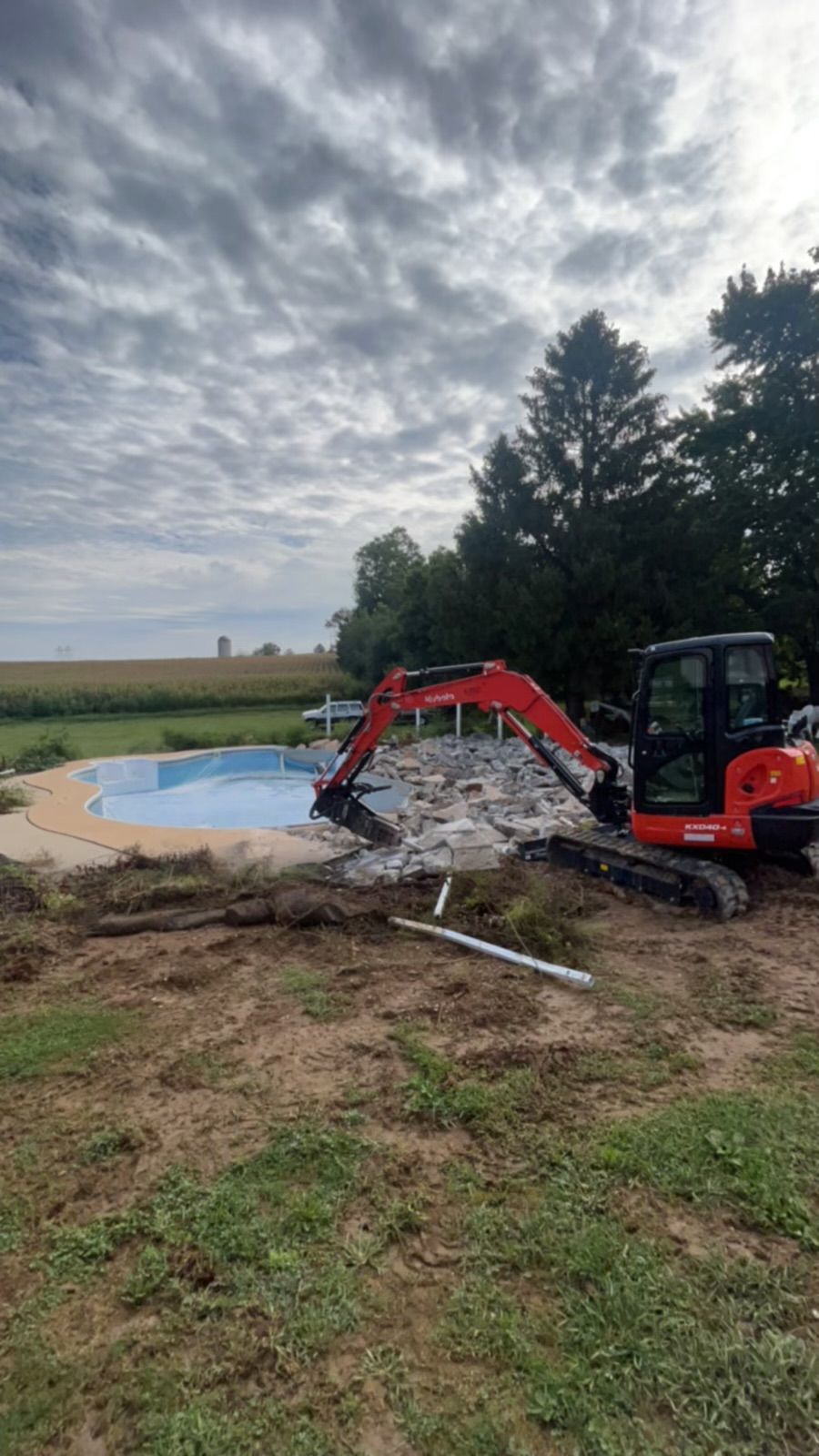 Excavator demolishing a concrete pool on a cloudy day, field in the background.