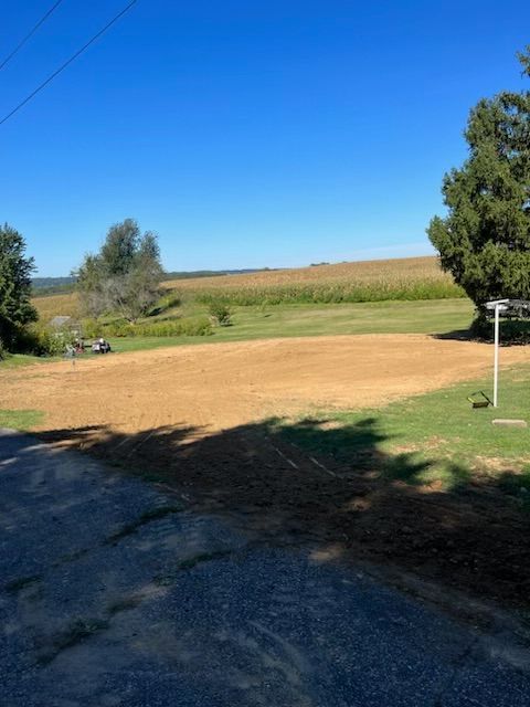 Sunny rural scene: cleared brown field, green grass, cornfield in the distance, blue sky.