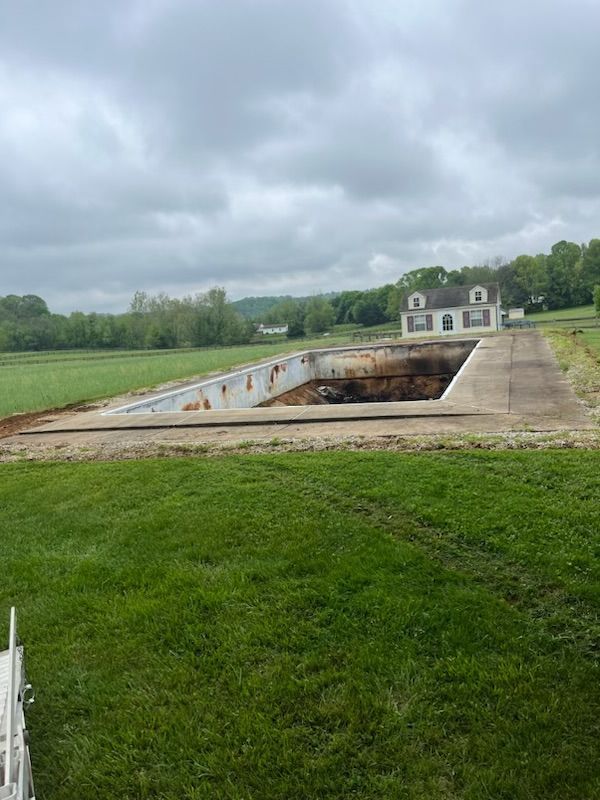 A collapsed rectangular pool in a grassy field with a house in the background under an overcast sky.