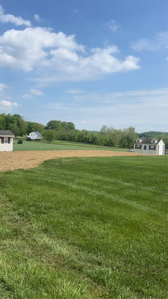 Green field with two white buildings, brown plowed section, trees, and a blue sky with clouds.