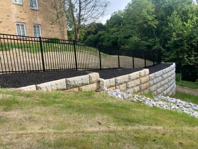 Black metal fence atop a stacked stone retaining wall with mulch, grass, and trees.