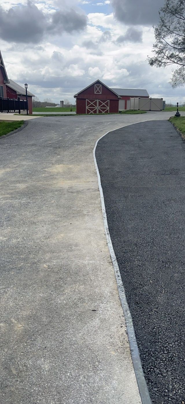 A gravel driveway curves towards red barns under a cloudy sky.