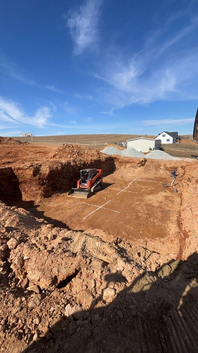 Excavated foundation pit with skid steer, red soil, and a clear blue sky.