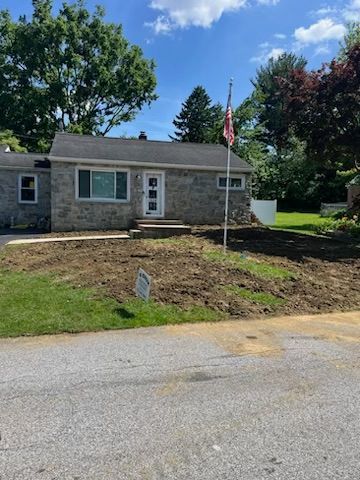 A one-story stone house with a partially cleared yard, flag, and a paved road.
