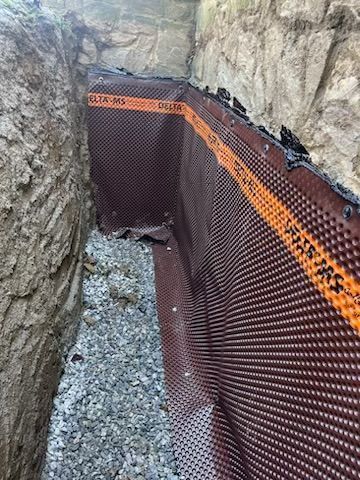 Dark brown dimpled drainage membrane lining a trench, with gravel base and dirt walls.