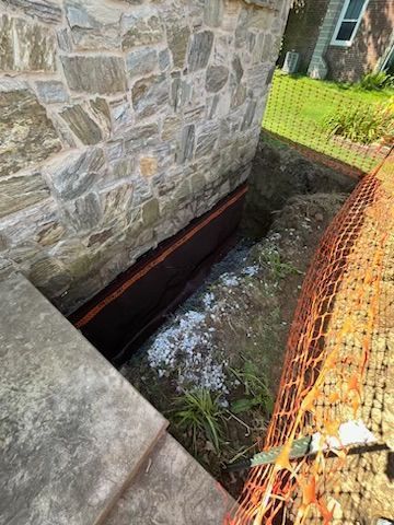 Trench dug beside a stone wall foundation, orange safety net visible, concrete stairs in foreground.