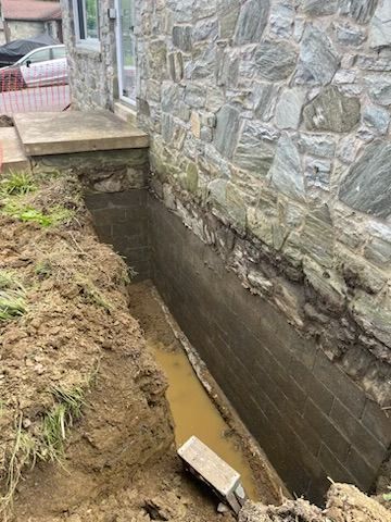 Exterior foundation repair: a trench dug beside a stone wall; a section of the foundation is exposed and coated.