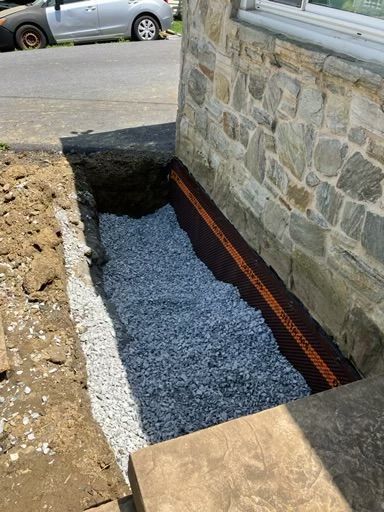 Trench next to a stone building foundation, filled with gravel and a drain, ready for backfill.