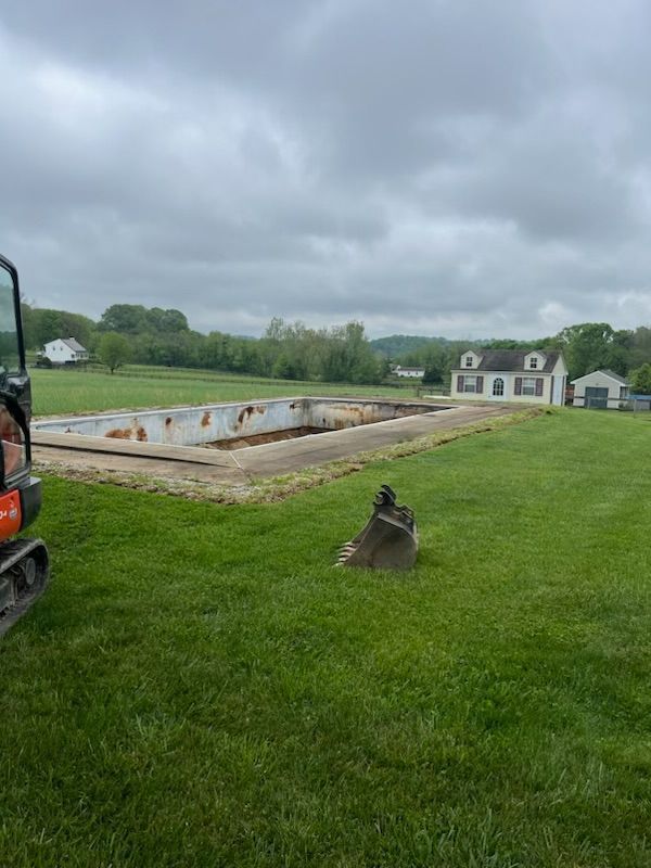 Excavator beside a rectangular concrete pool, partially demolished, on a grassy lawn with a house in the background. Overcast sky.