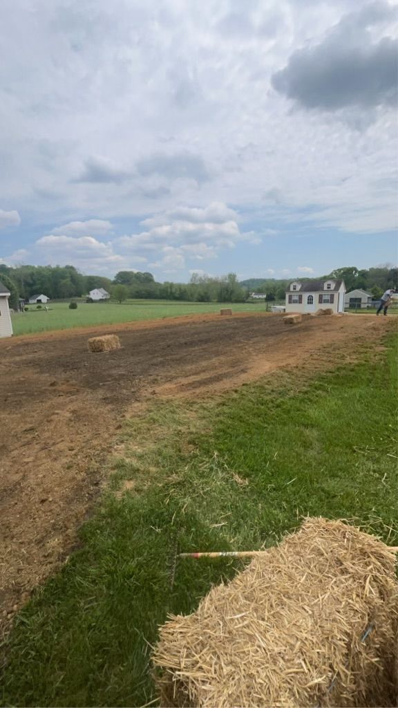 Grassy field with hay bales in foreground, tilled earth, small white house, trees, and cloudy sky.