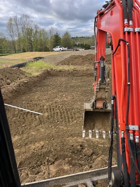An excavator digging in a dirt lot. The machine is orange. In the background is a truck. Cloudy sky.