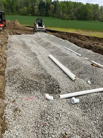 Construction site with gravel-filled trenches and white pipes. A small excavator and a skid steer are visible.