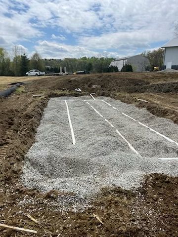Construction site with gravel-filled trench and white pipes for a septic system, outdoors under a cloudy sky.