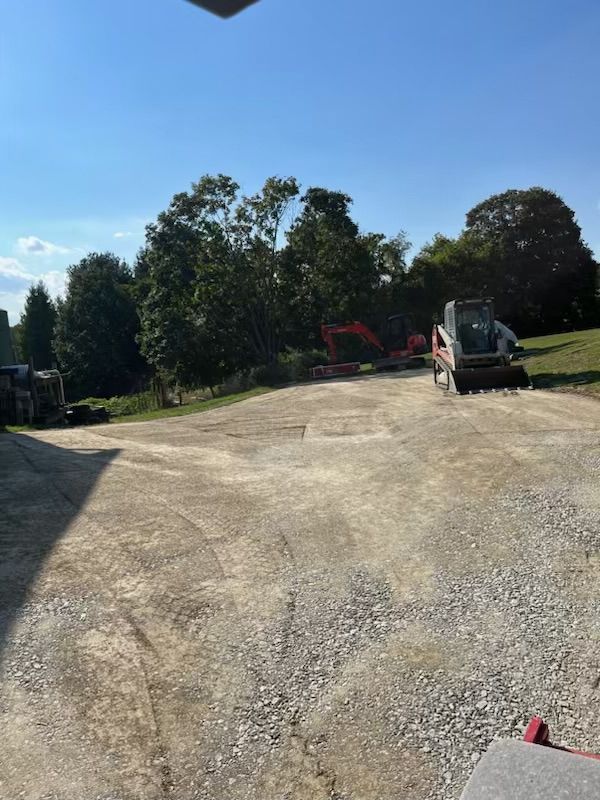Gravel driveway with construction equipment: Bobcat and excavator, with trees in the background under a blue sky.