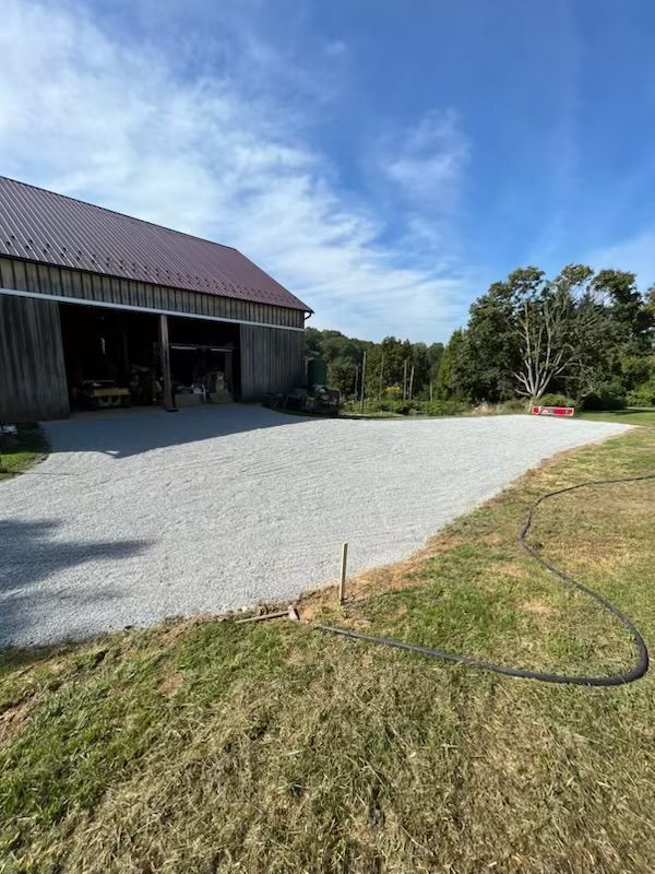 Gravel driveway leading to a wooden barn, set on a green grassy property under a blue sky.