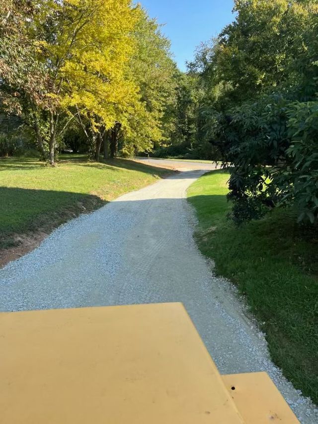 Gravel driveway curves through grassy yard with trees, seen from the top of yellow machinery.