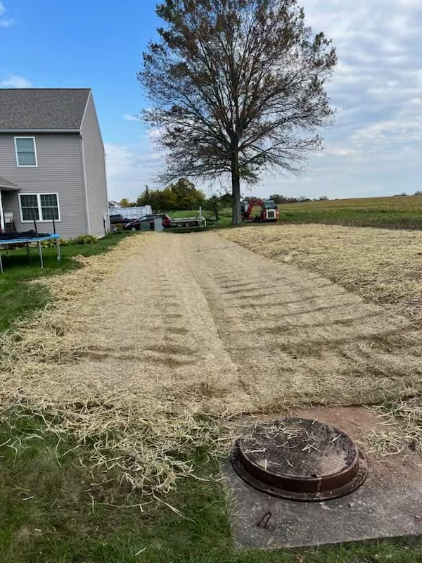 A residential backyard with gravel paths, a metal lid, and a house. A single tree is in the distance.