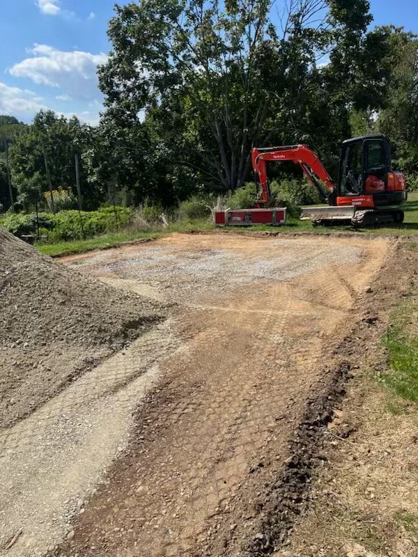 A construction site with a red excavator on a gravel pad, soil pile, and trees in the background.