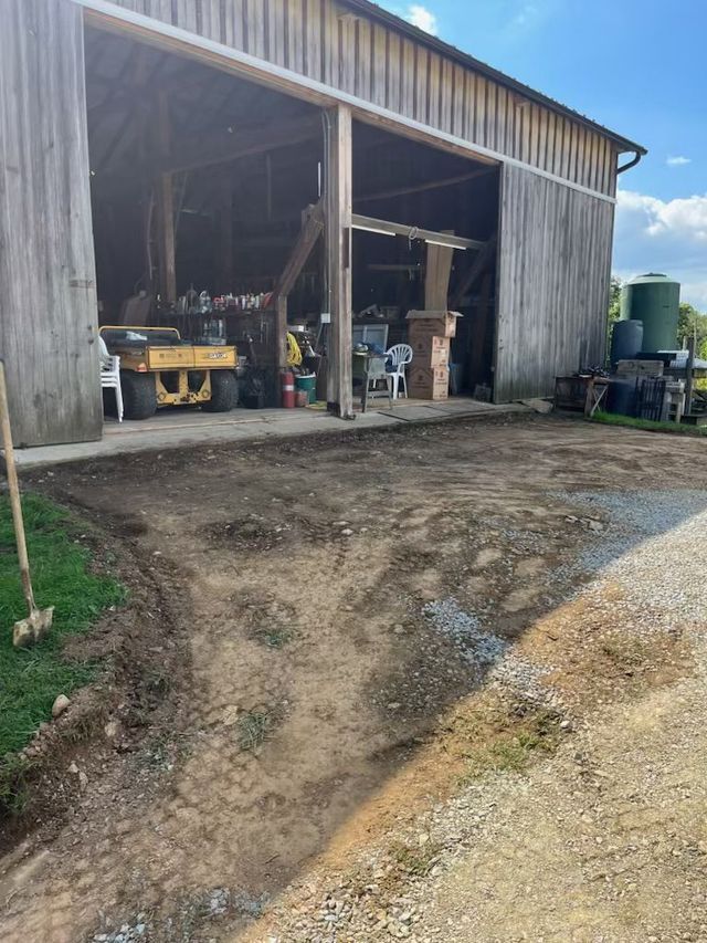 Barn with open doors, gravel driveway, and dirt ground. A yellow tractor is inside, and blue sky in background.