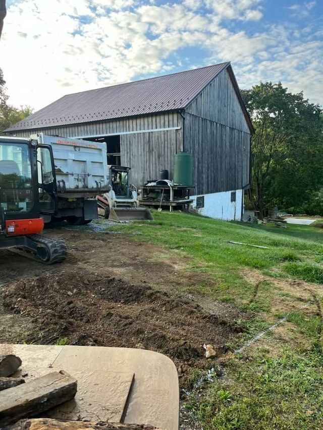 Barn with metal roof, excavator, and truck on a grassy hillside under a partly cloudy sky.
