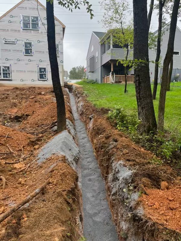 Long narrow trench filled with gravel, next to trees. Construction site with two houses in the background.