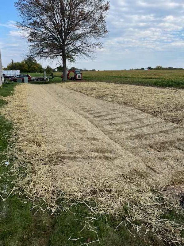 Field covered in light-colored straw, tractor in the distance, blue sky, and a few trees.