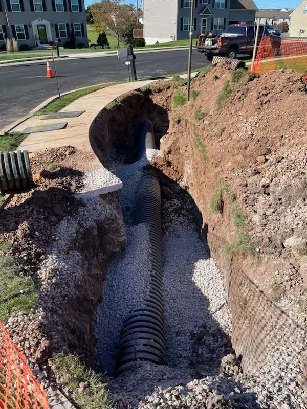 Construction site with trench revealing corrugated pipe surrounded by gravel. Sidewalk and street in the background.