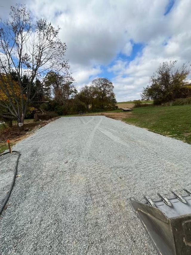 Gravel driveway under a cloudy sky, leading to a green field with trees.