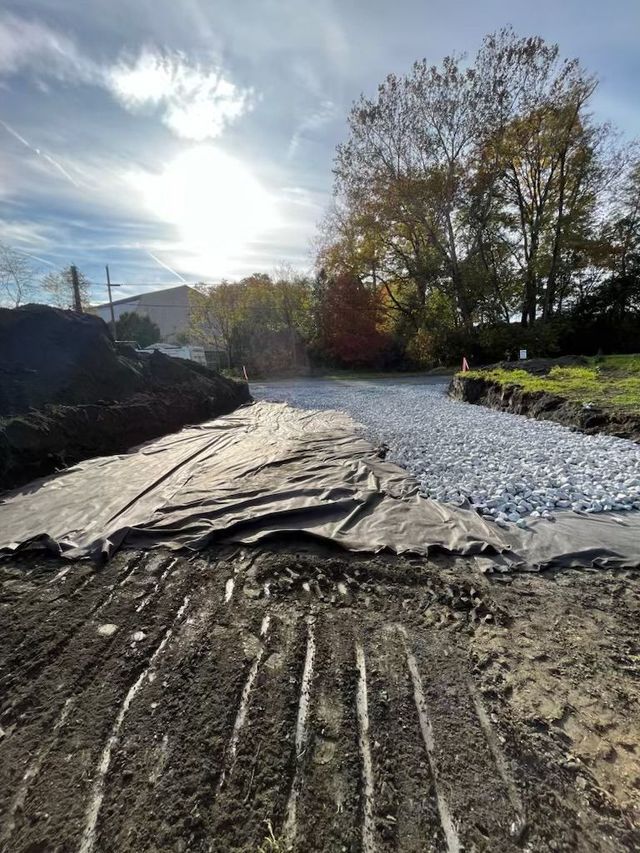 Construction site: ground graded, covered with fabric, and filled with gravel; trees and sky in background.