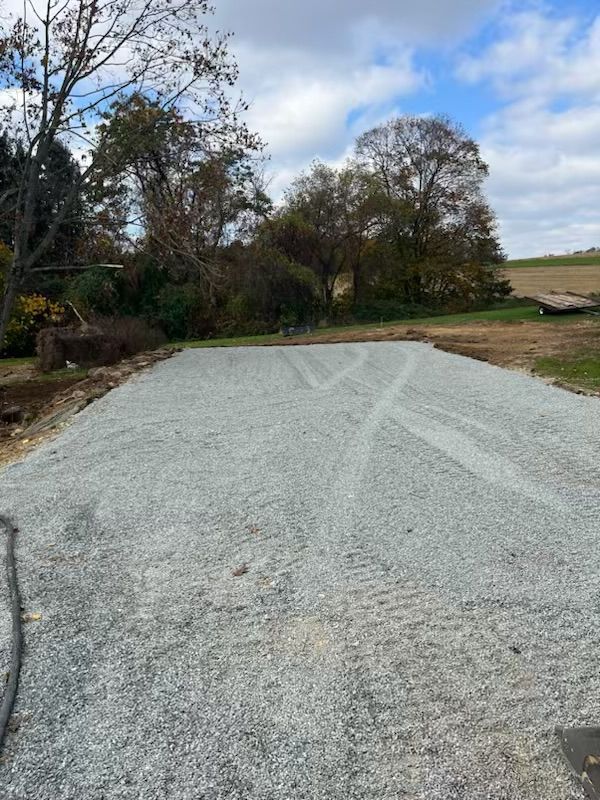 Gravel driveway in a grassy area with trees in the background under a cloudy sky.