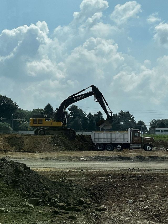 Yellow excavator loading dirt into a brown dump truck at a construction site under a cloudy sky.