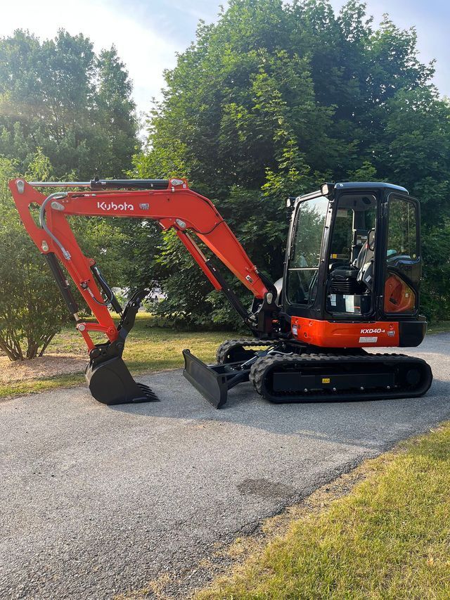 Orange and black Kubota excavator on a paved driveway in front of trees.