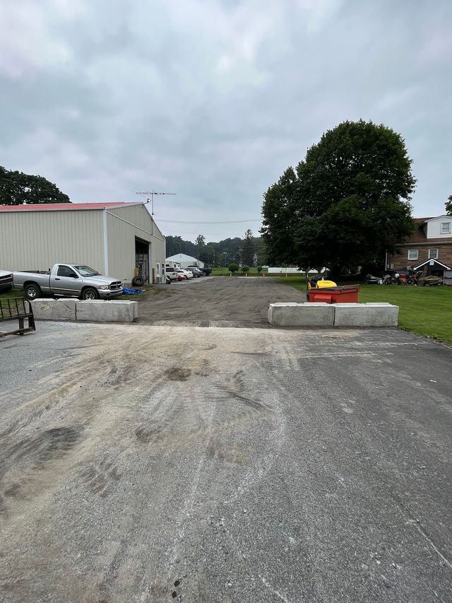 Paved road blocked by concrete barriers, leading to industrial buildings. Cloudy sky overhead.