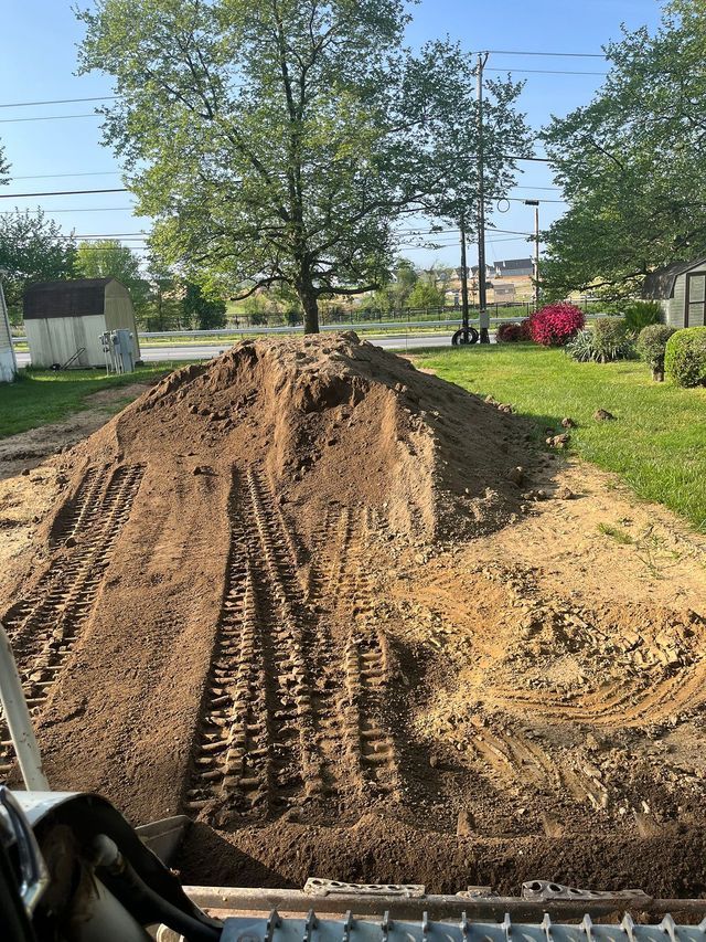 Large pile of dirt with track marks on a grassy yard, trees in the background, blue sky.