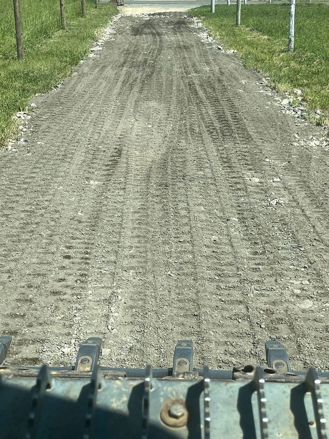 Gravel driveway with tire tracks, viewed from inside machinery. Green grass borders on both sides.