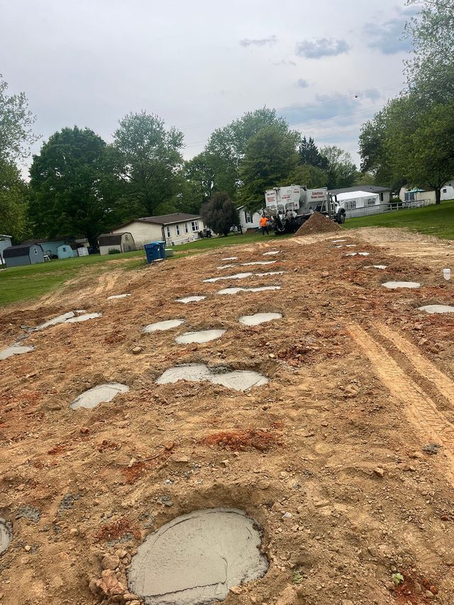 Construction site with numerous circular concrete pads in dirt. Background includes trees and homes.