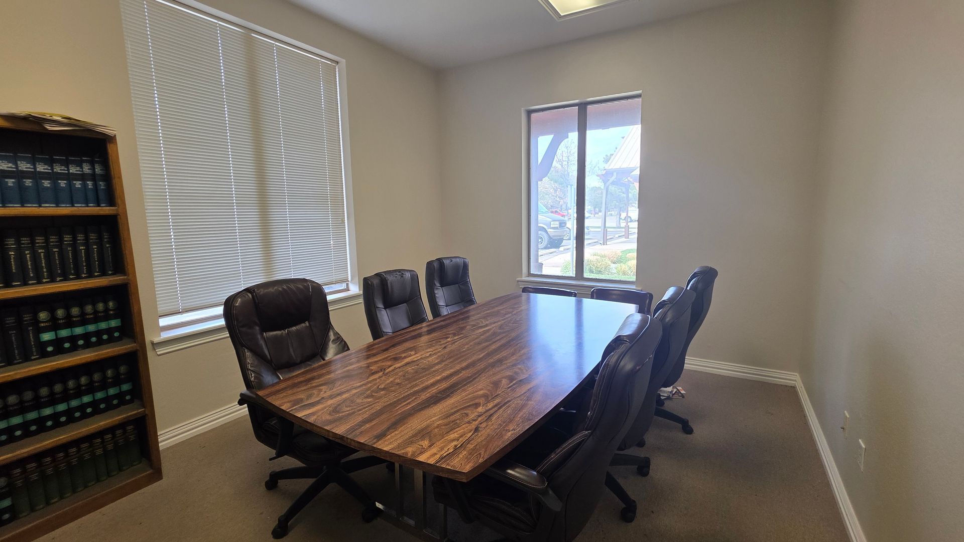 A conference room featuring a long wooden table, six black leather office chairs, and a tall bookshelf filled with books.