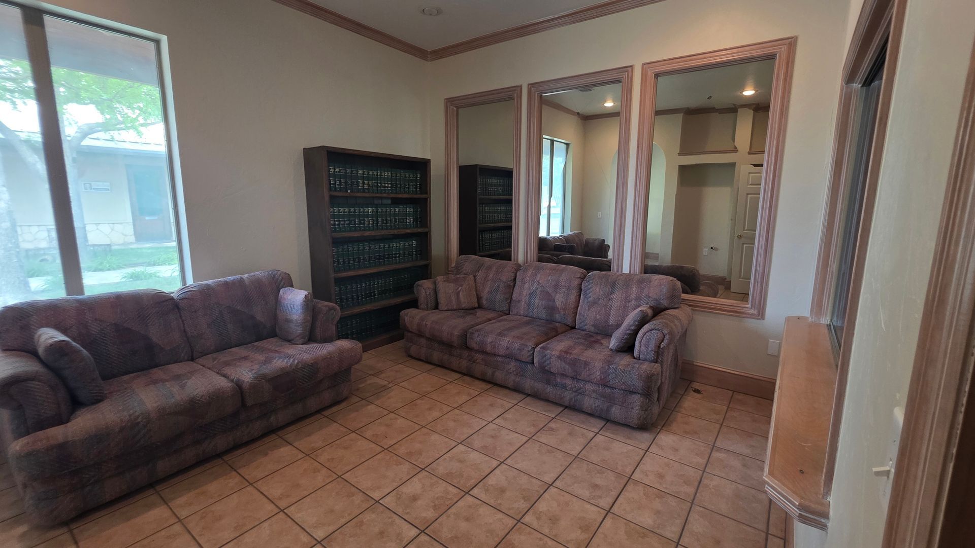 A room featuring two brown fabric sofas, a tall bookshelf, and large wall mirrors, with tile flooring.