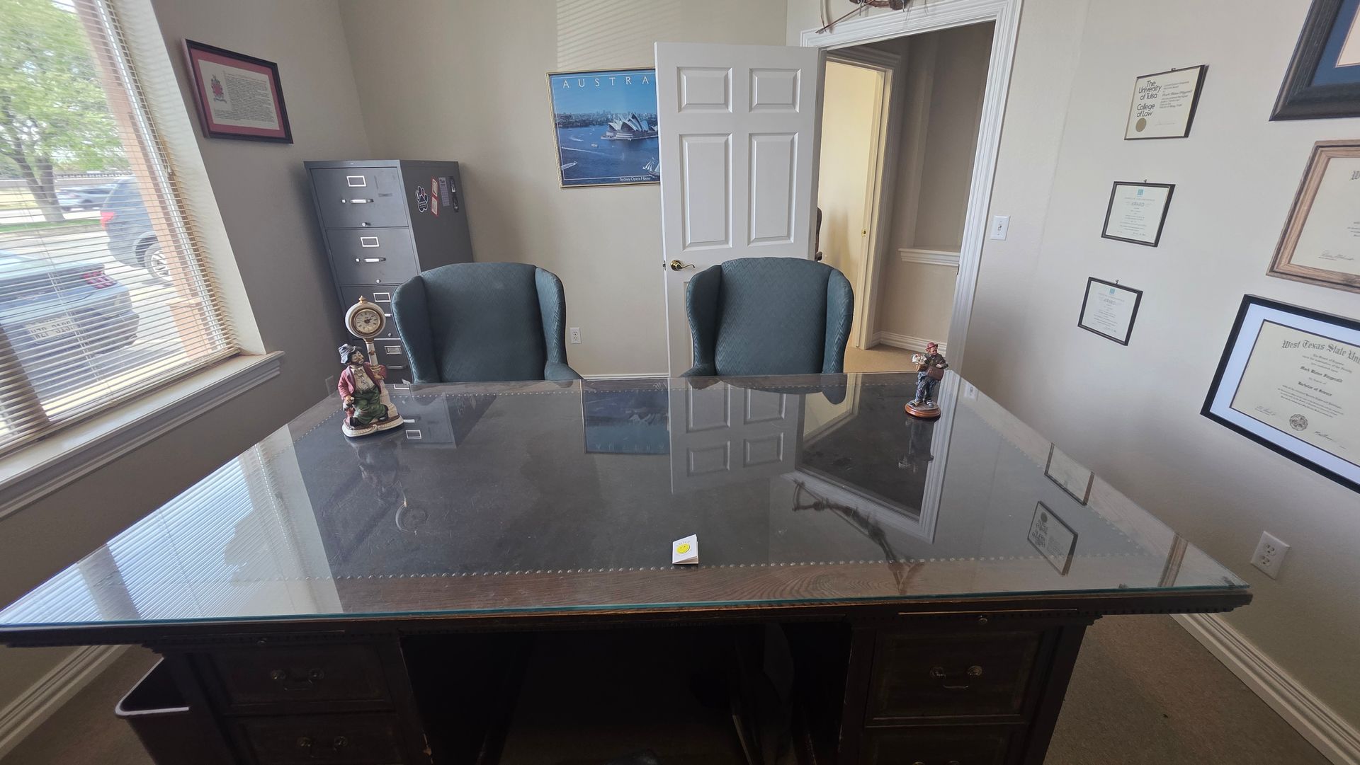 A dark wood desk with a glass top sits in a professional office, featuring two dark chairs and framed wall certificates.