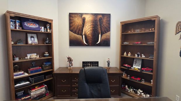 A home office with a dark wood desk, central chair, and a large elephant painting flanked by two shelves of collectibles.