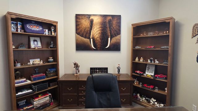 A home office with a dark wood desk, central chair, and a large elephant painting flanked by two shelves of collectibles.