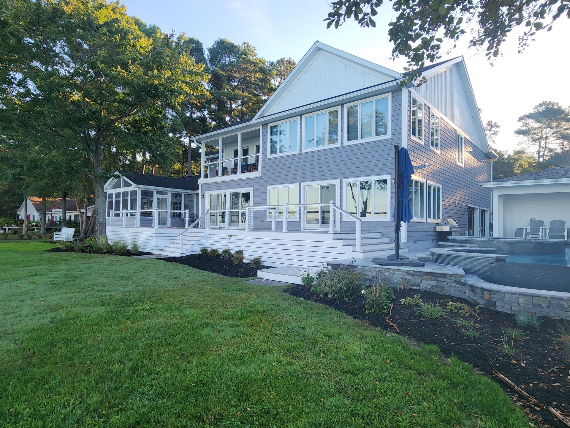 Two-story gray house with white trim and deck, on a green lawn. Blue umbrella and hot tub visible.