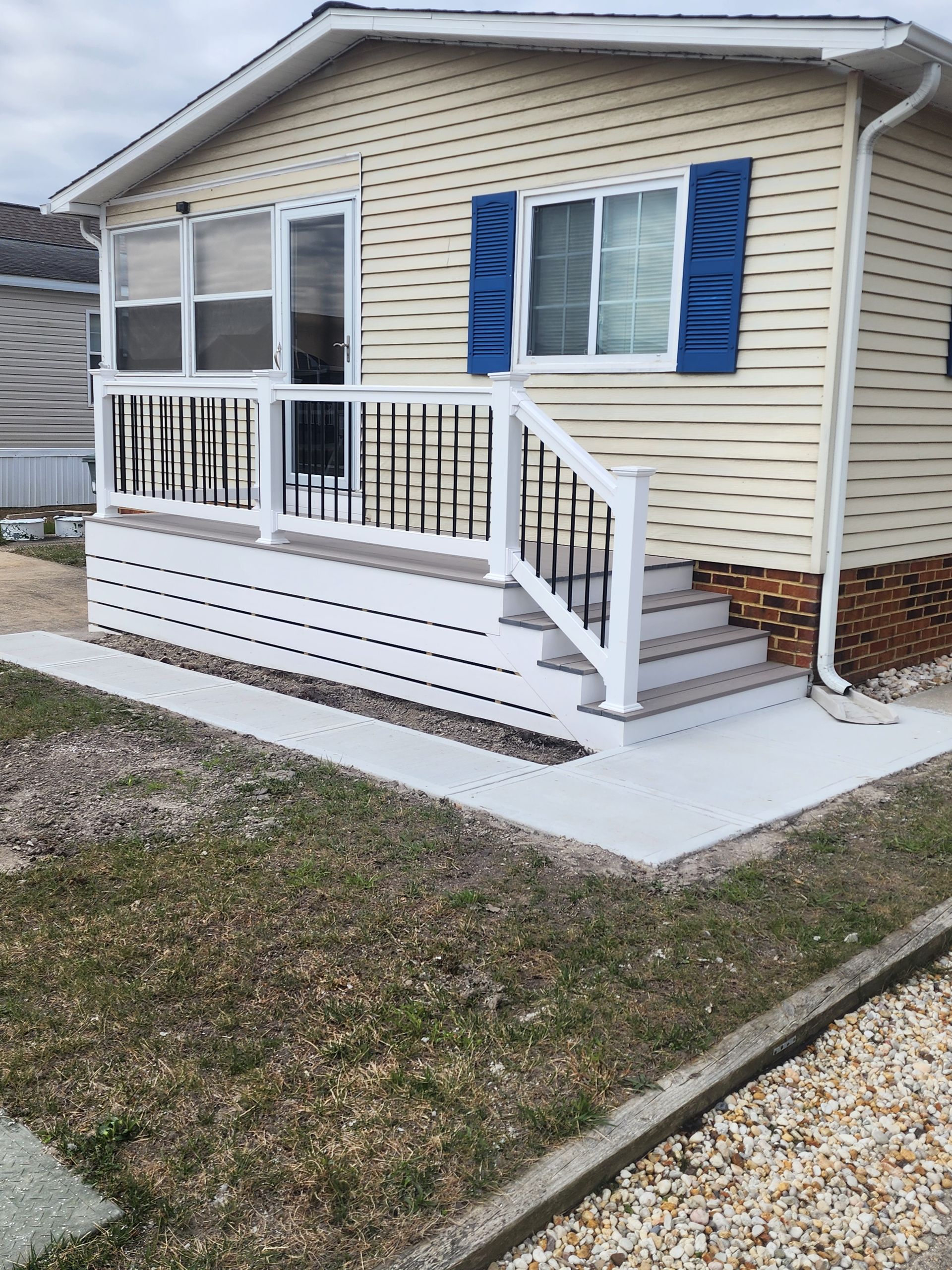 A small beige house with a white porch and blue shutters. Concrete path and steps lead up to the entrance.