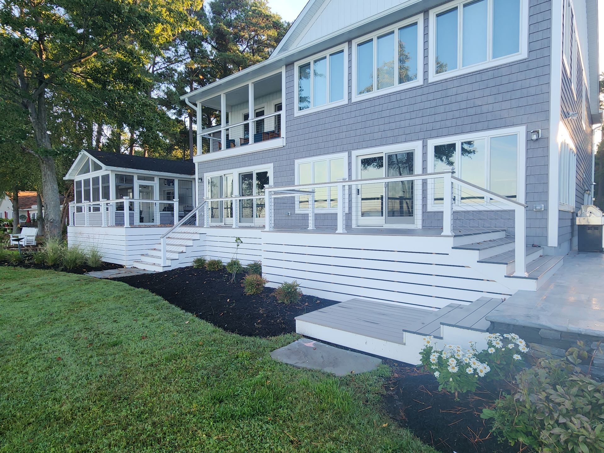 Two-story house with white deck, glass railings, and a screened porch overlooking a grassy lawn.