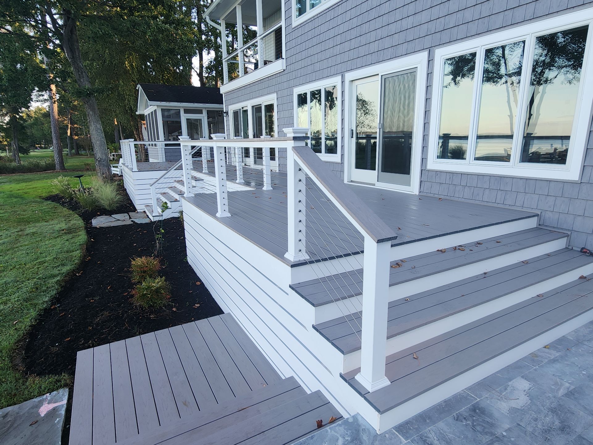 Gray deck with white railing and steps leading to a house with large windows; landscaping in foreground.