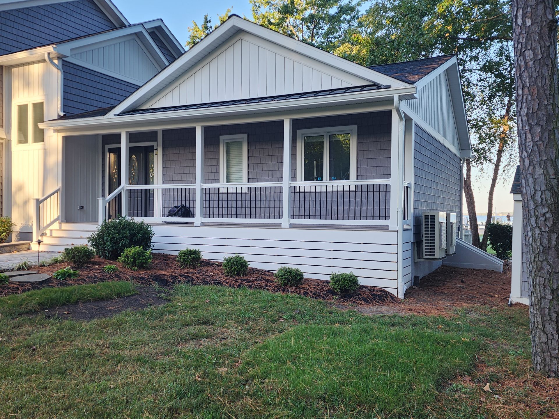 Gray house with white porch, black railings, and side paneling. Lush green lawn in front.