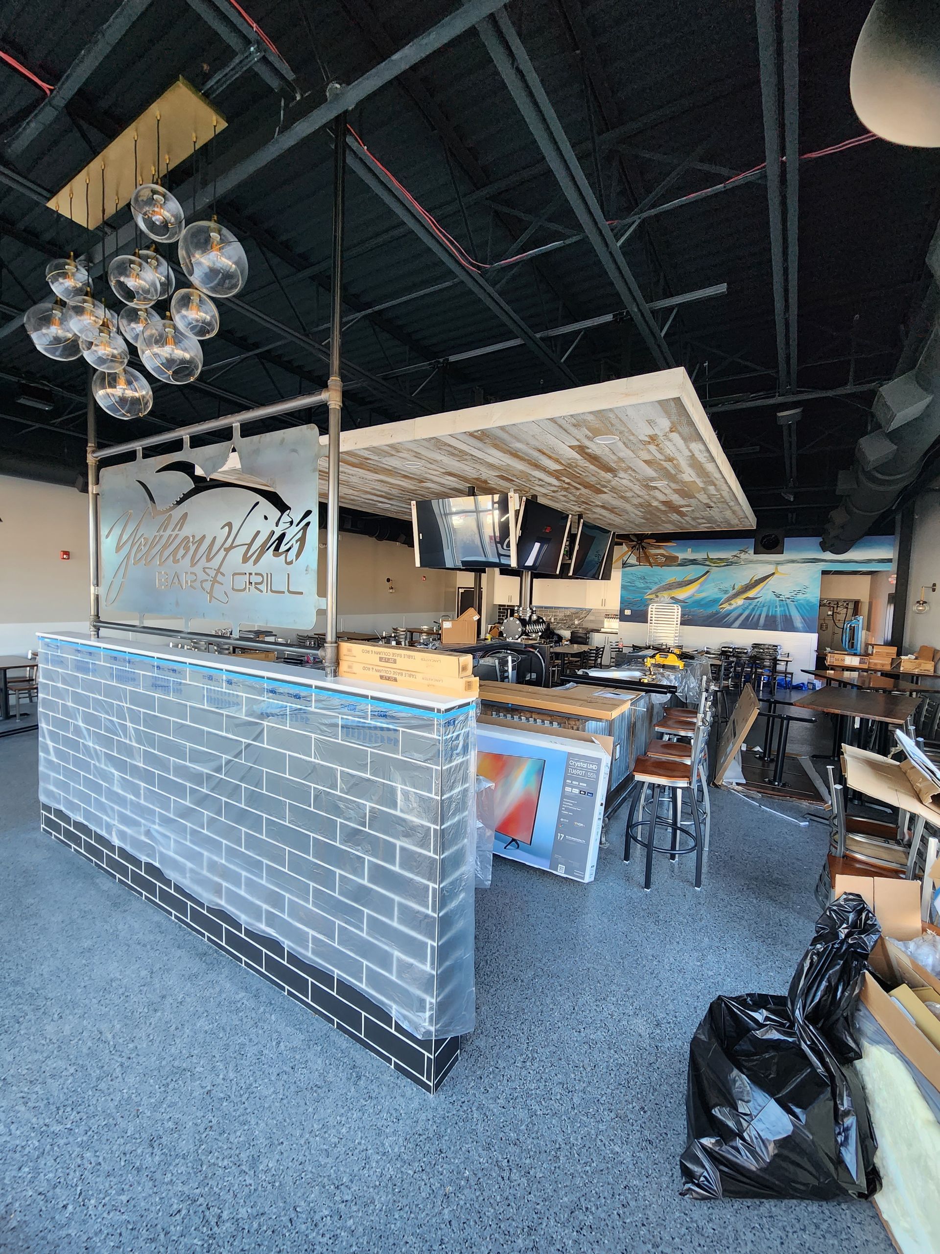 Interior of a restaurant. Bar with brick facade, metal stools, and overhead lighting. Gray speckled floor.