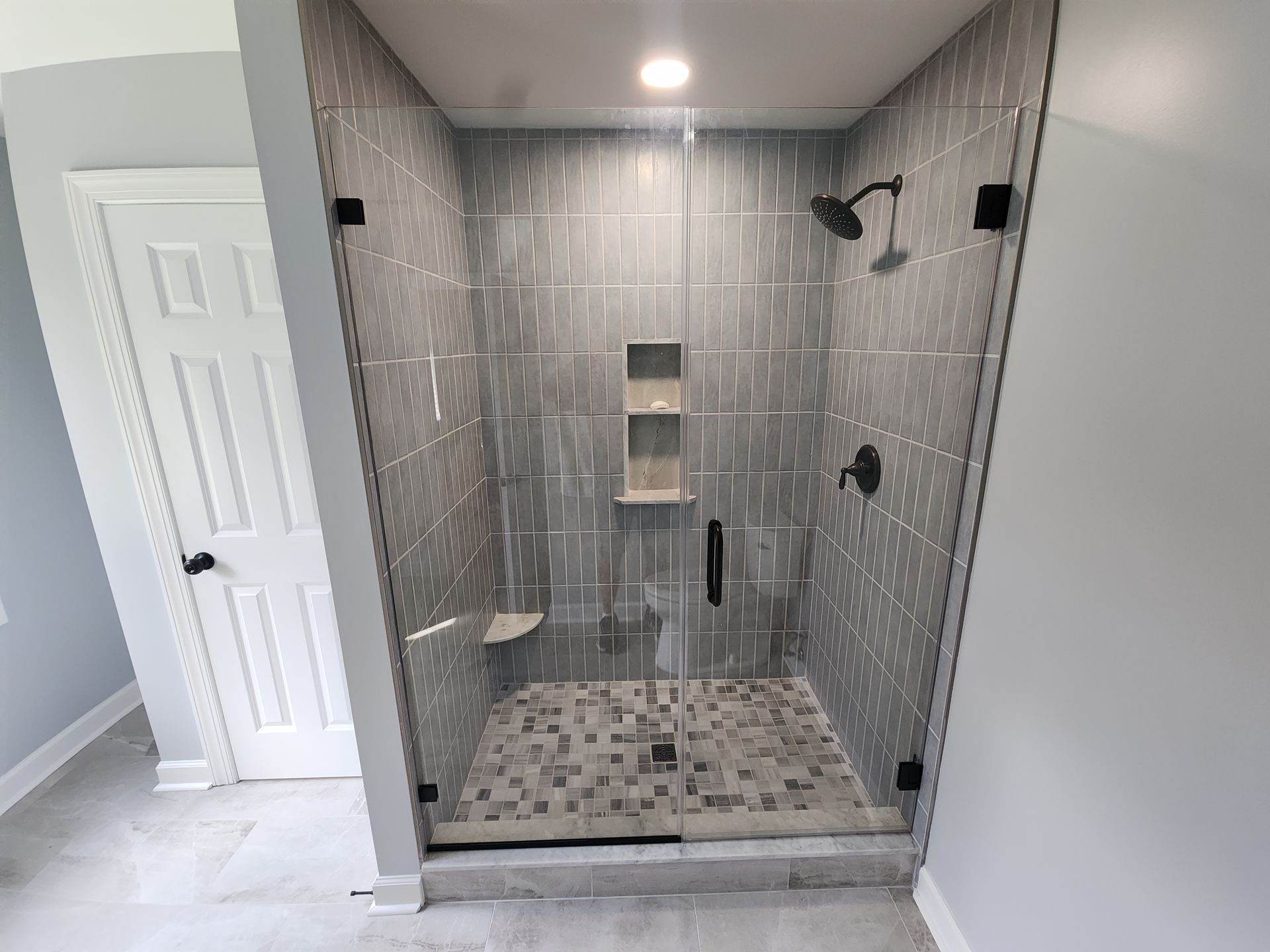 Gray tiled shower with glass doors and black fixtures; a white door is visible on the left.