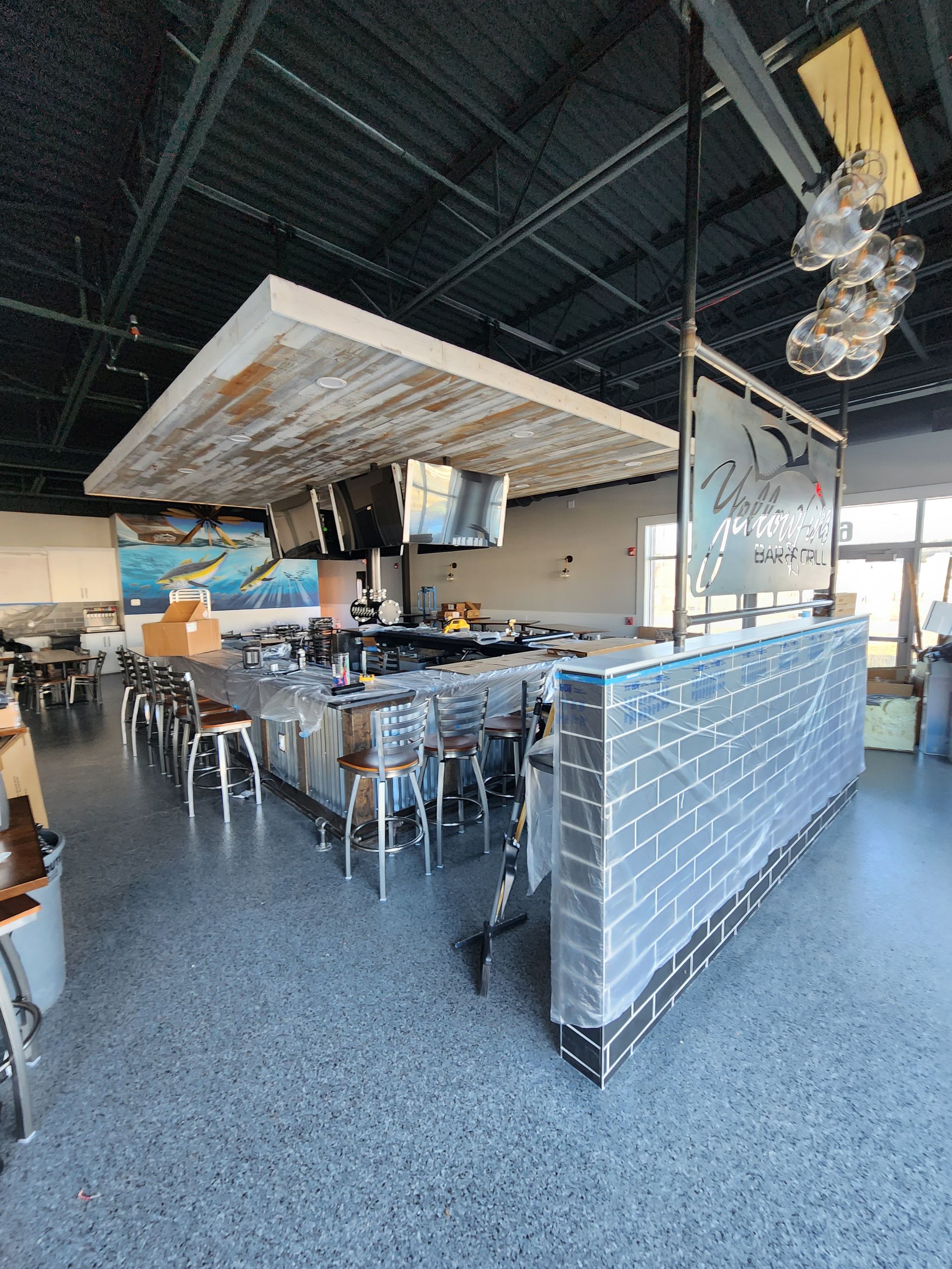 Bar with stainless steel counter, metal stools, brick facade, and TVs. Light fixture and artwork visible.